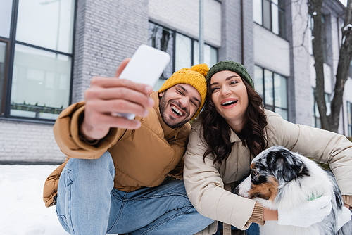smiling man in knitted hat taking selfie with girlfriend and australian shepherd dog in winter