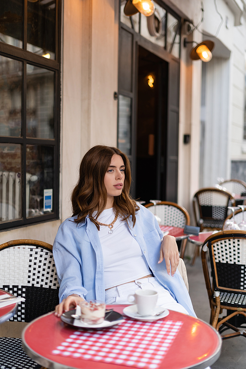 Stylish woman sitting near blurred coffee and dessert on table in outdoor cafe in Paris