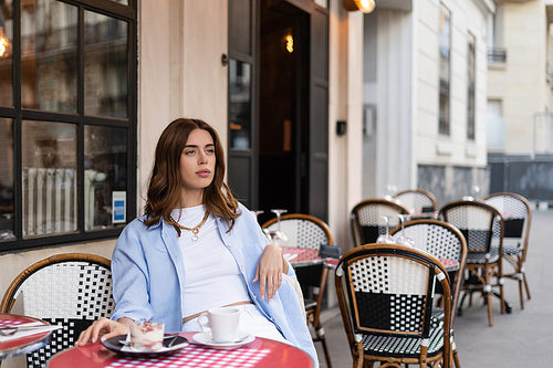 Stylish woman looking away while sitting near dessert and coffee on table on cafe terrace in Paris