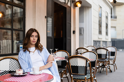 Brunette woman looking at camera near coffee and dessert in outdoor cafe in Paris