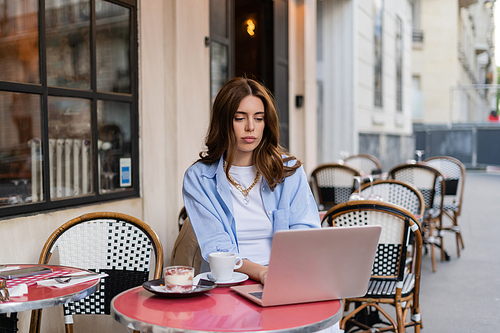 Stylish freelancer using laptop near coffee and dessert on cafe terrace in Paris