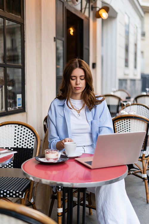 Freelancer holding coffee cup near dessert and laptop on table of cafe in Paris