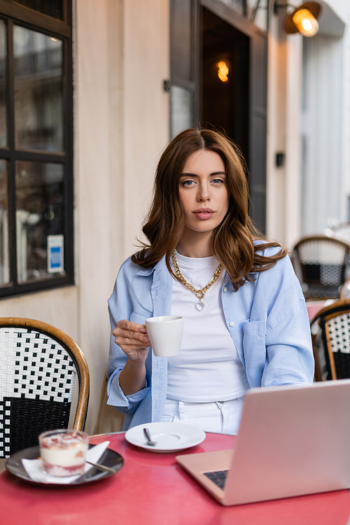Stylish woman holding cup of coffee near blurred dessert and laptop in outdoor cafe in Paris