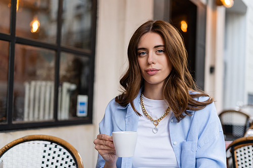 Stylish brunette woman holding cup of coffee on cafe terrace in Paris