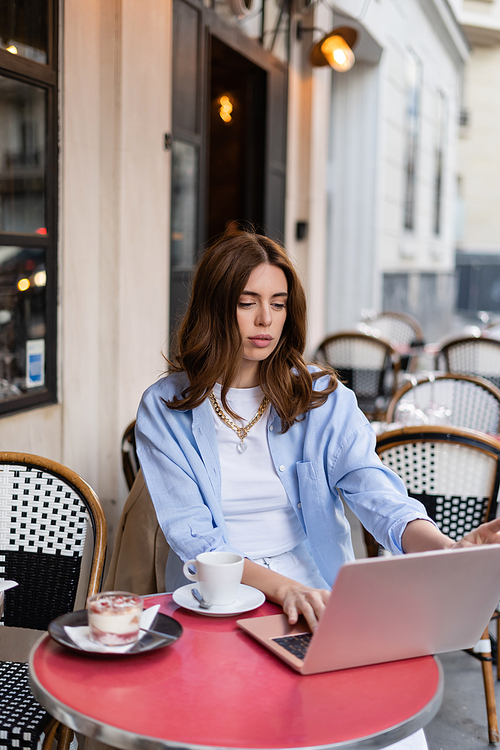 Trendy freelancer using laptop near coffee and dessert in outdoor cafe in Paris