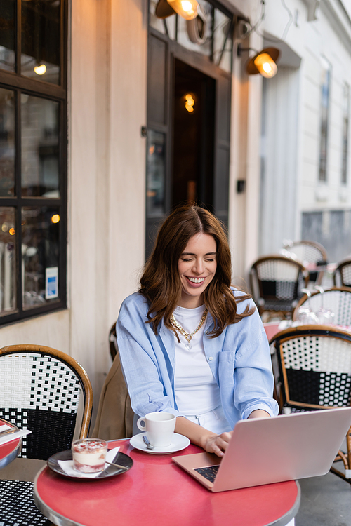 Cheerful freelancer using laptop near dessert and coffee in outdoor cafe in France