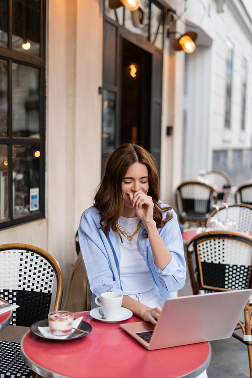 Positive freelancer using laptop near coffee and dessert in cafe on urban street in France