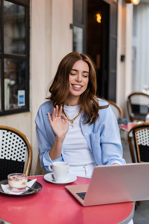 Stylish woman having video call on laptop near coffee and desert in outdoor cafe in Paris