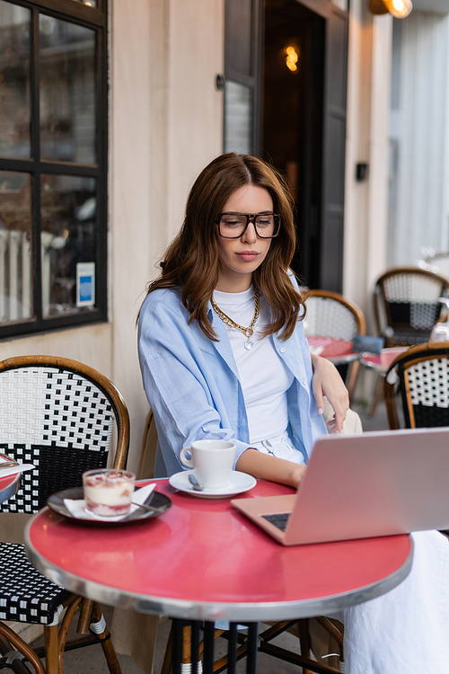 Stylish freelancer using laptop near dessert and coffee in outdoor cafe in Paris