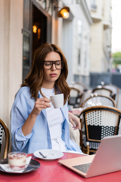 Stylish woman holding cup near laptop and blurred dessert on cafe terrace in Paris