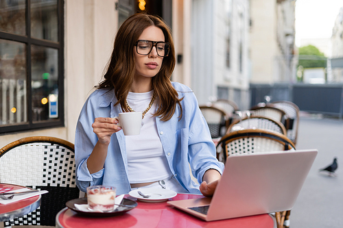 Trendy freelancer holding cup near laptop and dessert in outdoor cafe in Paris