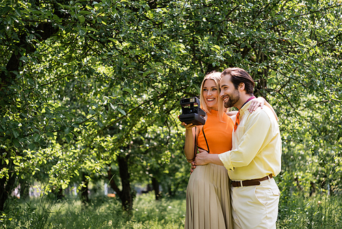 Smiling man hugging stylish girlfriend with vintage camera in summer park