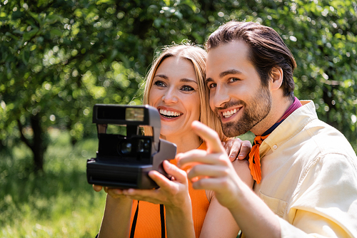 Stylish man pointing with finger near girlfriend with blurred retro camera in park