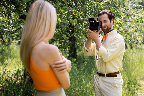 Trendy man taking photo on vintage camera near blurred girlfriend in summer park