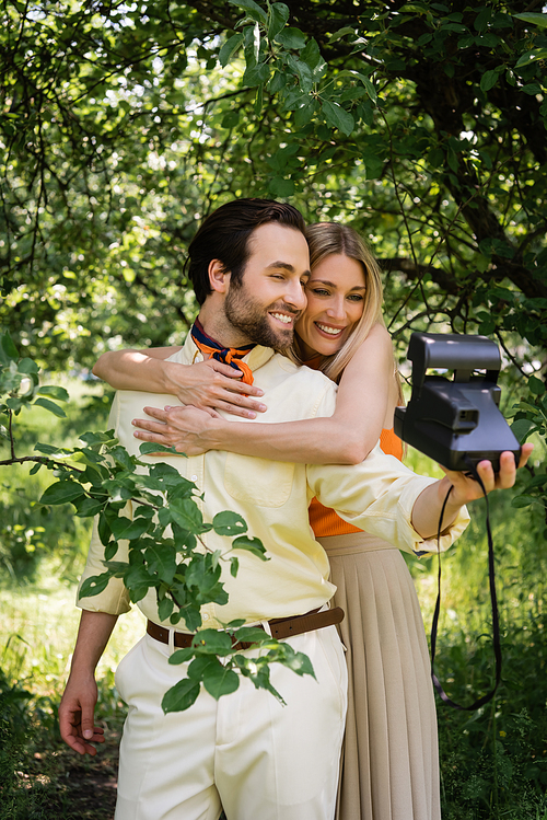 Smiling woman embracing stylish boyfriend taking photo on retro camera in summer park