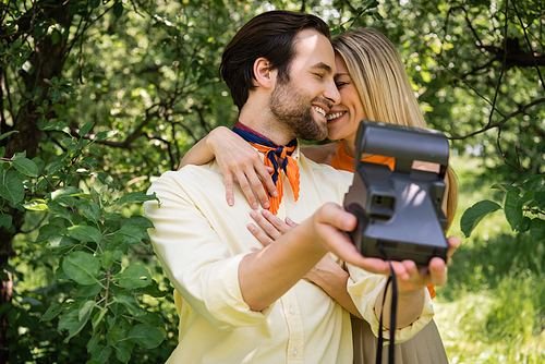 Cheerful woman embracing stylish boyfriend with blurred retro camera in park