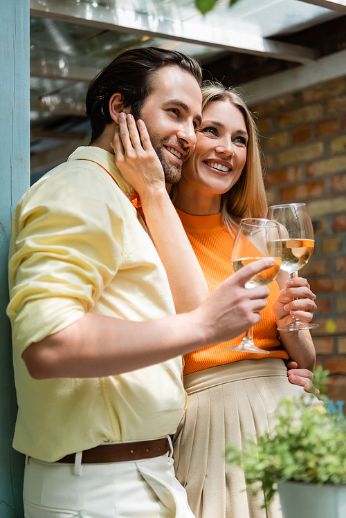 Stylish woman touching face of smiling boyfriend with wine in outdoor cafe