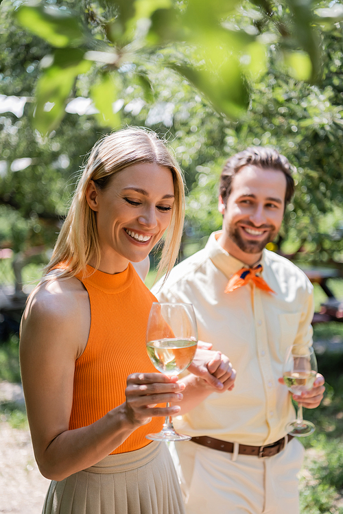 Cheerful woman holding glass of wine near blurred boyfriend in summer park