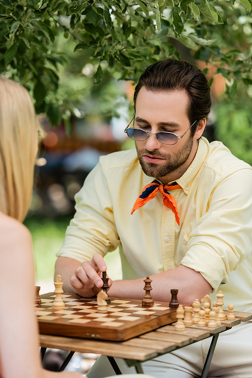 Stylish man in sunglasses playing chess with blurred woman in summer park