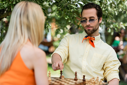 Trendy young man pointing at chess figure near blurred girlfriend in summer park