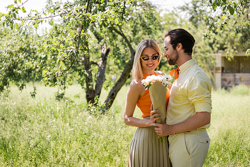 Fashionable man in sunglasses holding bouquet near smiling girlfriend in summer park