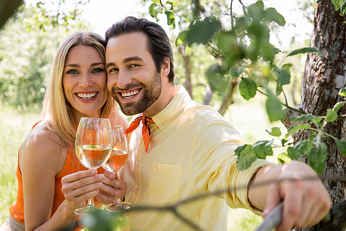 Smiling and trendy couple holding glasses of wine near tree in summer park