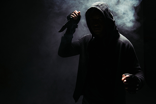 Silhouette of african american bandit in hoodie holding knife on black background with smoke