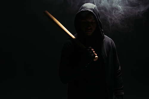 Silhouette of african american hooligan in hood holding baseball bat on black background with smoke