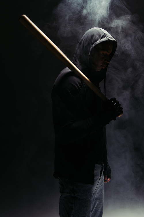 Side view of silhouette of african american bandit holding baseball bat on black background with smoke