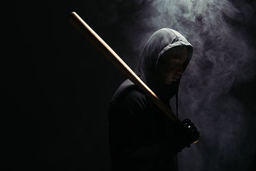 Side view of african american hooligan holding baseball bat in lighting on black background with smoke