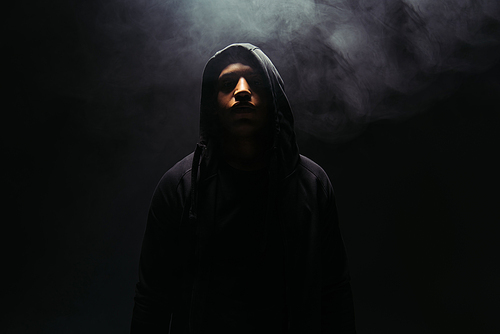 African american hooligan in hoodie standing on black background with smoke