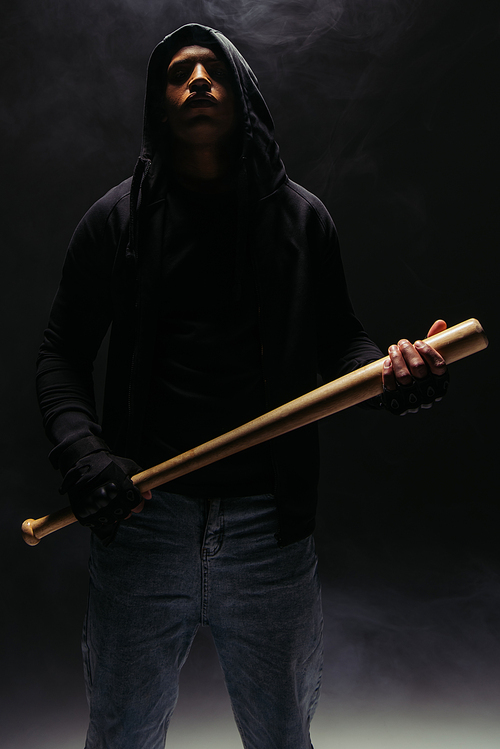 African american bandit in hood holding baseball bat on black background with smoke