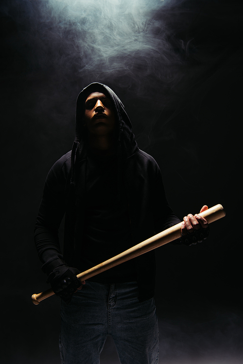 African american hooligan in hoodie holding baseball bat on black background with lighting and smoke