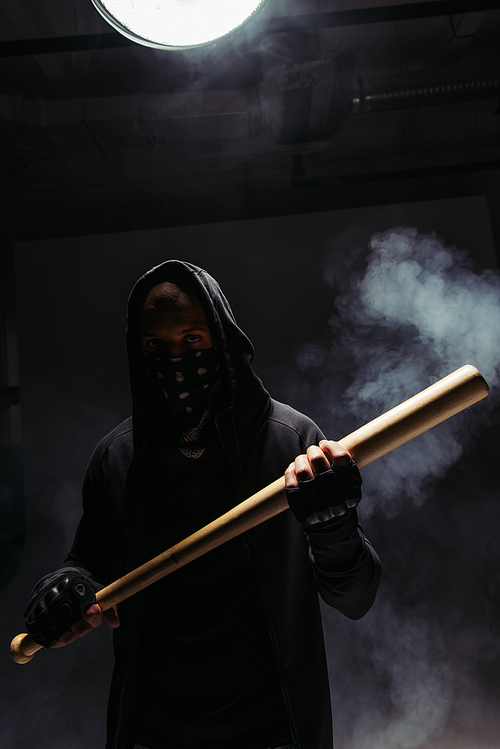 Silhouette of african american vandal with mask on face holding baseball bat on black background with smoke