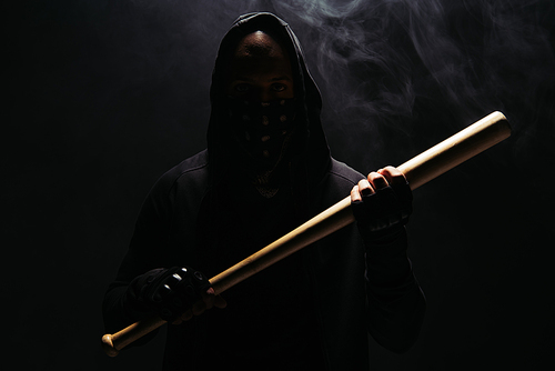 Silhouette of african american man in hoodie and gloves holding baseball bat on black background with smoke