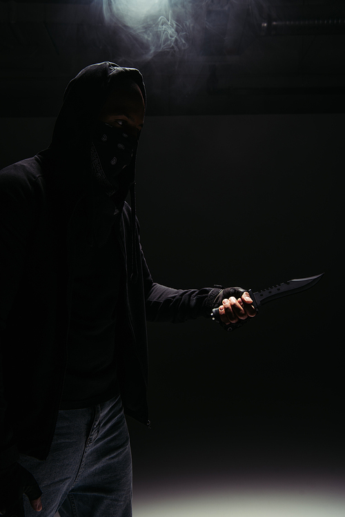 Silhouette of african american robber in mask and hoodie holding knife on black background with smoke