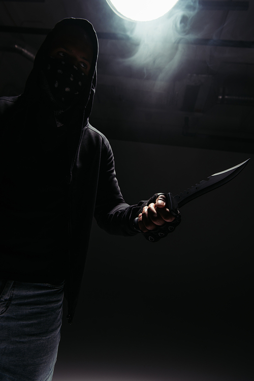 Silhouette of african american hooligan with obscure face holding knife on black background with smoke