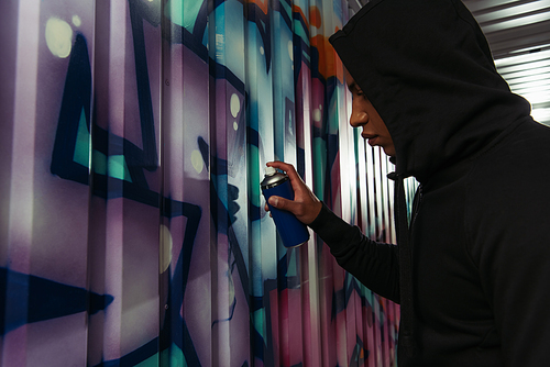 Side view of african american vandal in hood drawing graffiti on wall