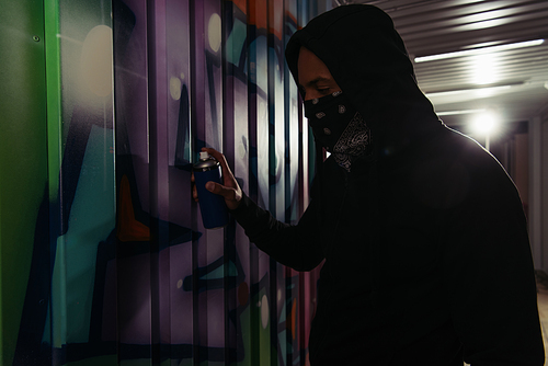 Side view of african american hooligan in hoodie and mask drawing graffiti on wall