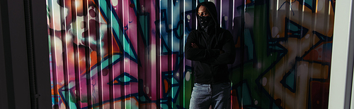 African american hooligan in hoodie and face mask standing near graffiti on wall, banner