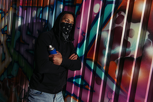 African american hooligan in hoodie holding spray paint near graffiti on wall on urban street