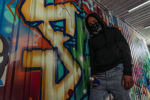 Low angle view of african american vandal with mask on face holding spray paint near graffiti