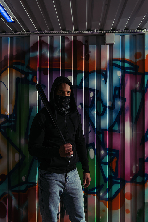 African american hooligan in mask holding baseball bat near graffiti on wall