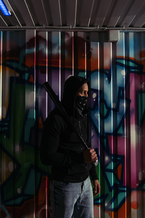 Side view of african american hooligan in mask and hoodie holding baseball bat near graffiti on wall