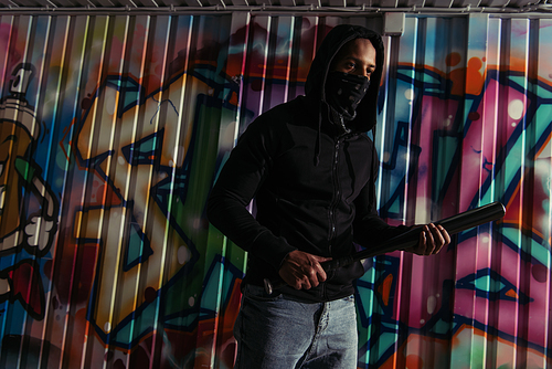 African american hooligan in hoodie holding baseball bat near graffiti on wall