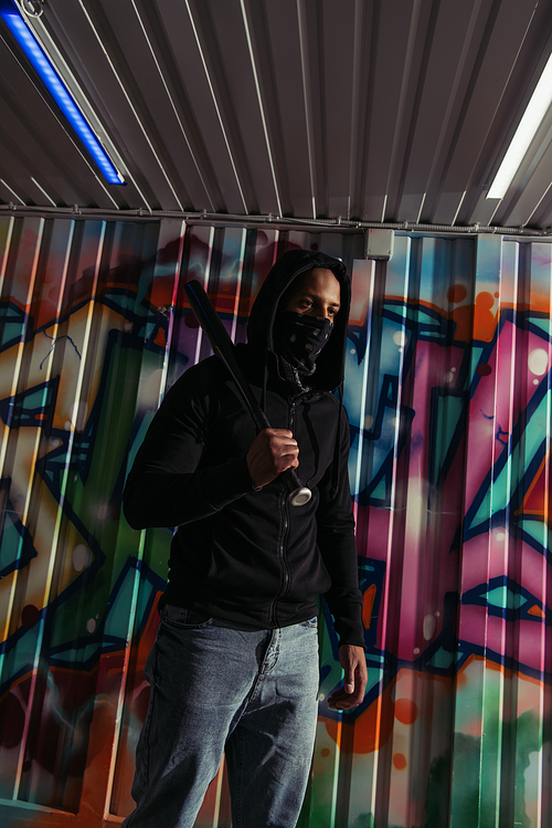 African american vandal in mask holding baseball bat near graffiti on wall