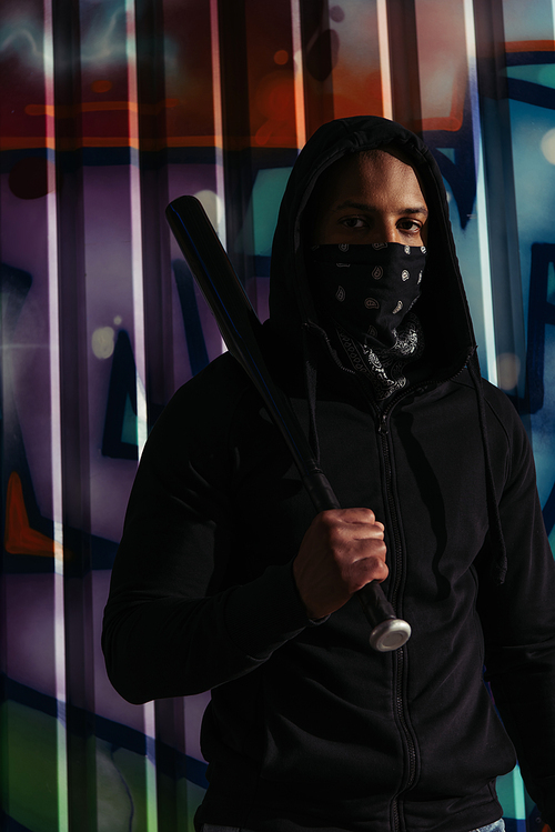 African american hooligan holding baseball bat and looking at camera near graffiti on urban street