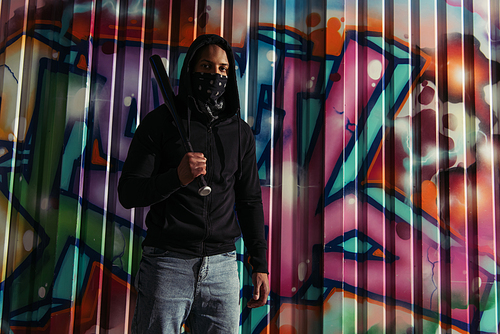 African american hooligan holding baseball bat and looking at camera near graffiti outdoors