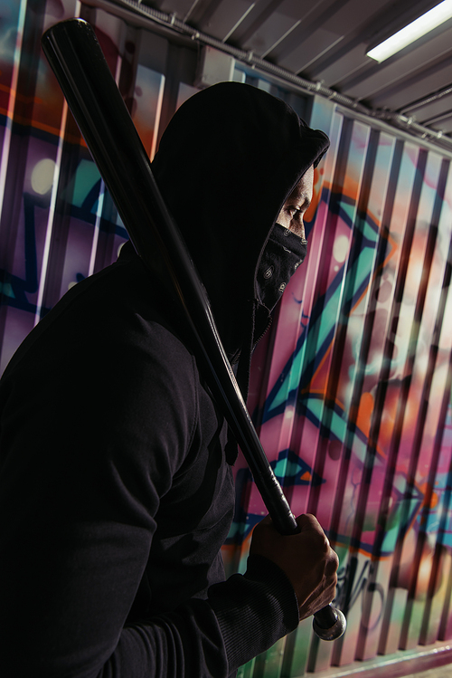 Side view of african american vandal in mask holding baseball bat near graffiti on wall