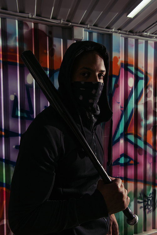 African american hooligan with baseball bat looking at camera near graffiti on wall